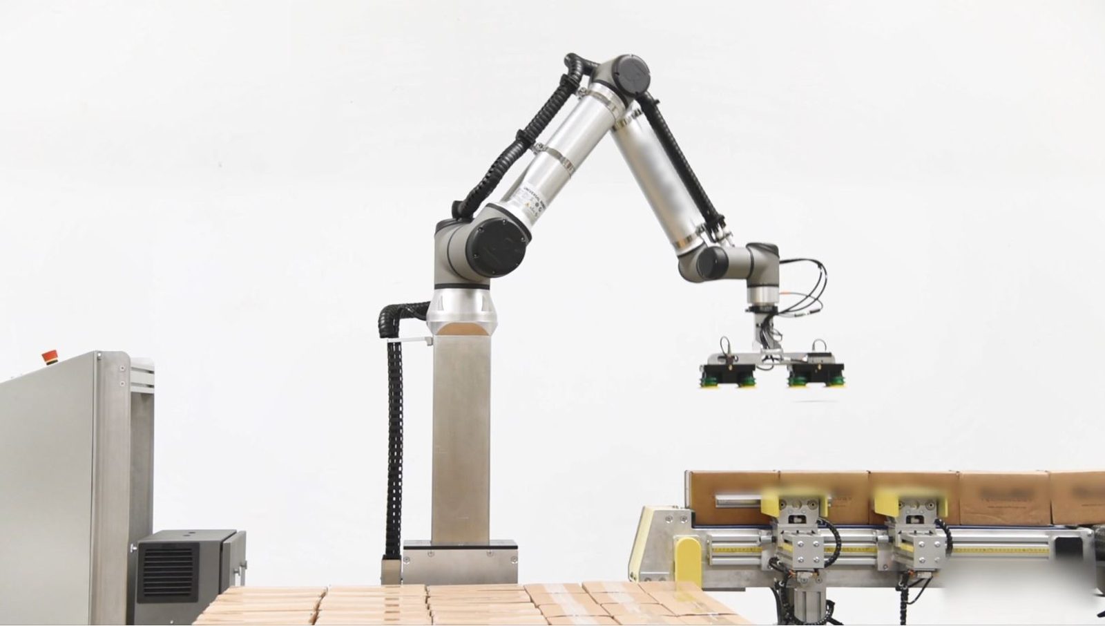 Universal Robots cobot arm mounted on a lift column, palletizing boxes onto a pallet in a compact fenceless palletizing cell.